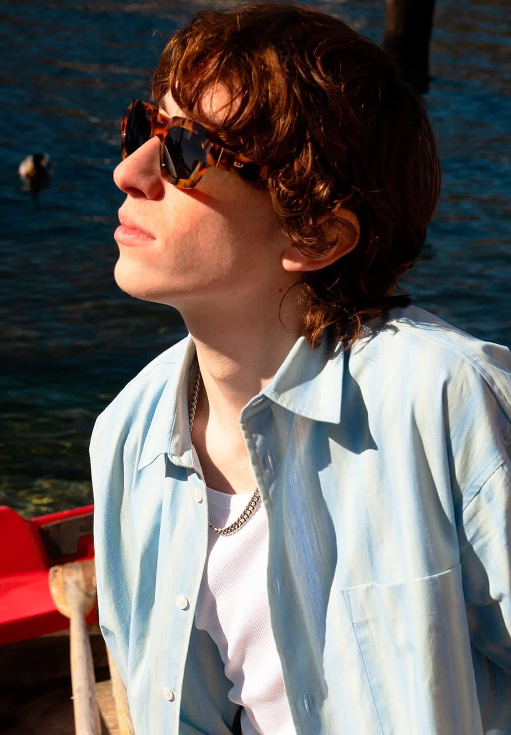 Occhiali da sole tartarugati, camicia azzurra chiara con finitura testurizzata e collana in argento. Ambientazione luminosa all'aperto vicino all'acqua.