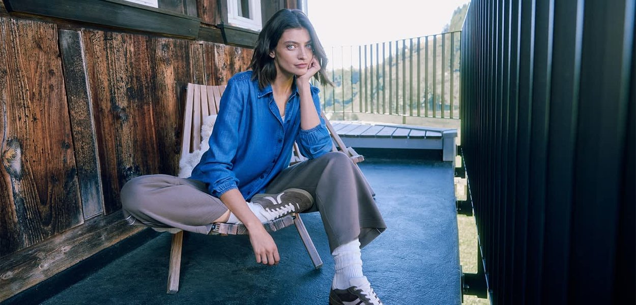 Fauteuil en bois sur un balcon, une personne portant une chemise en denim bleu, un pantalon gris et des baskets marron, assise les jambes croisées, au soleil.