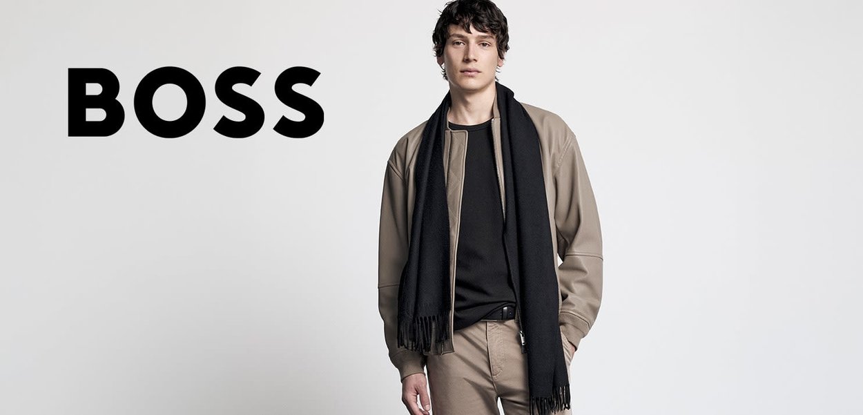 Beige Bomberjacke über einem schwarzen Hemd, kombiniert mit sandfarbenen Hosen. Ein langer schwarzer Schal hängt herab und hat Fransenakzente.