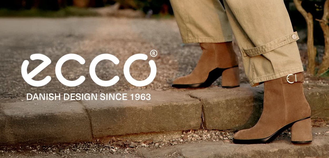 Brown suede ankle boots with a block heel and a side buckle detail, paired with light beige trousers, set against a stone path.