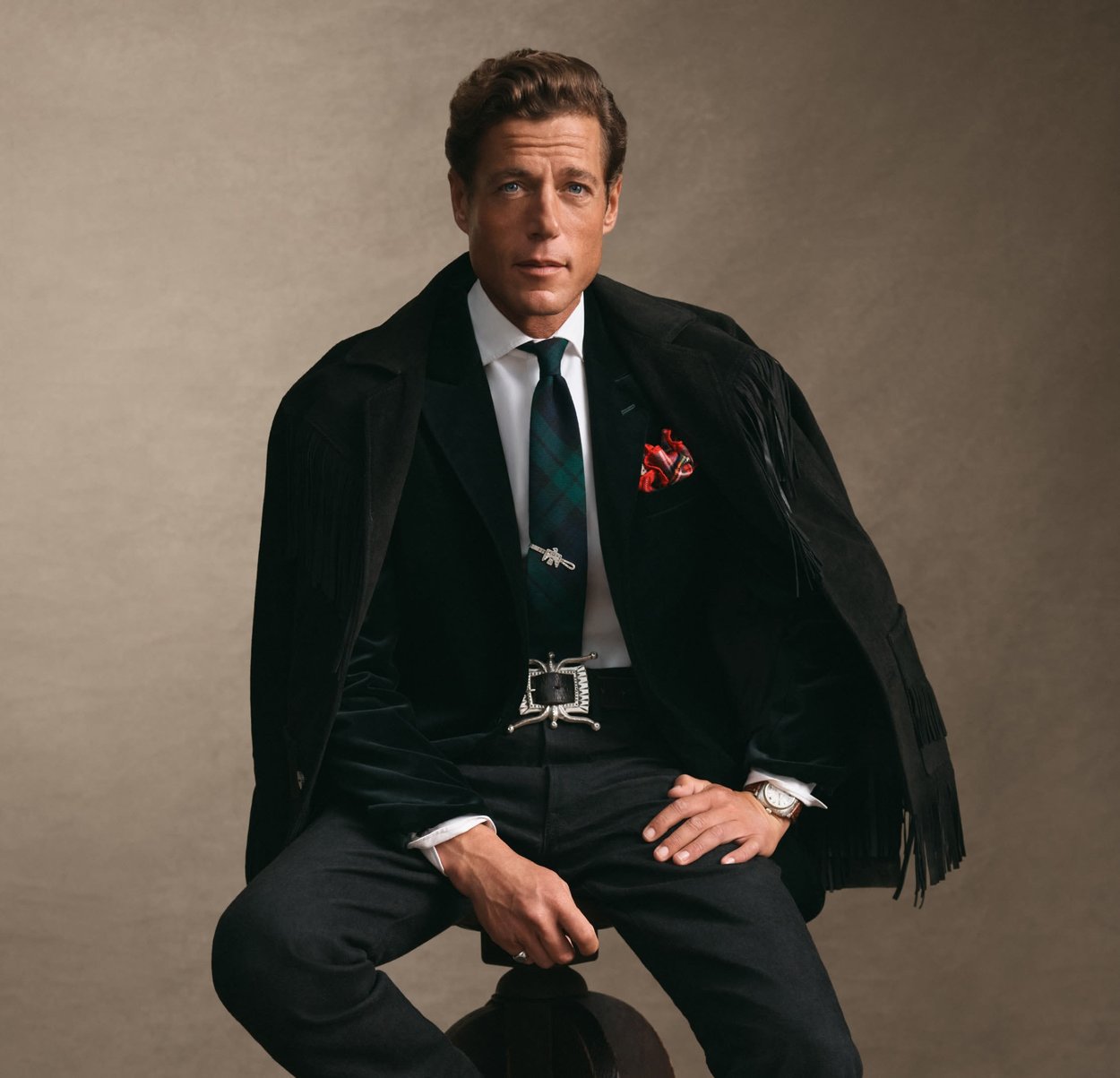 Homme assis sur un tabouret, portant une veste noire à franges sur une chemise blanche, une cravate à carreaux, un mouchoir rouge, une boucle de ceinture en argent et un pantalon sombre.