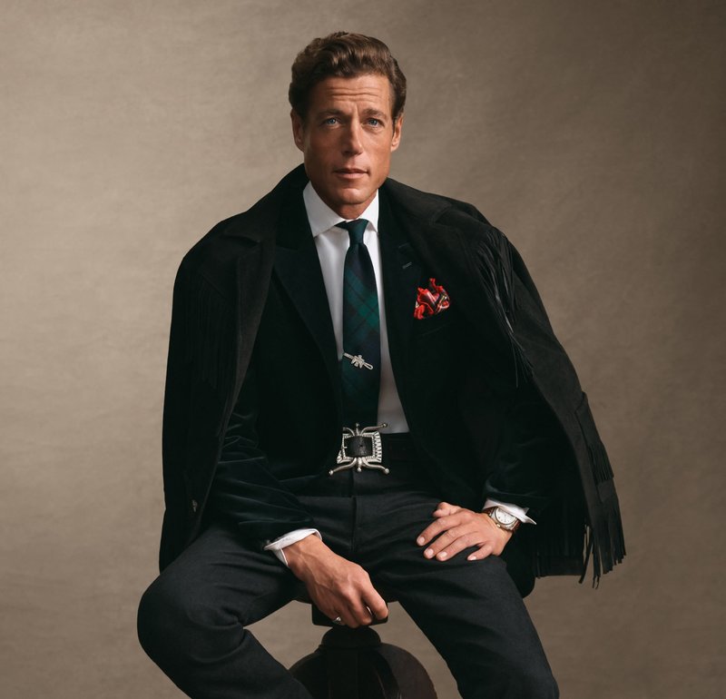 Homme assis sur un tabouret, portant une veste noire à franges sur une chemise blanche, une cravate à carreaux, un mouchoir rouge, une boucle de ceinture en argent et un pantalon sombre.