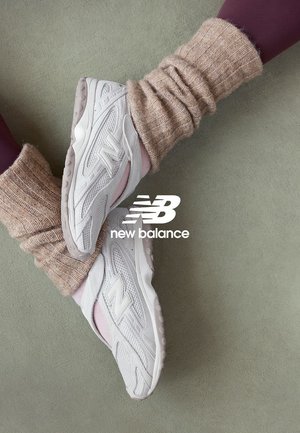 Zapatillas blancas New Balance combinadas con calentadores de piernas de punto beige y leggings morados sobre un fondo de alfombra verde claro.