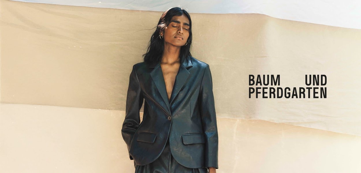 Kobieta z zamkniętymi oczami, ubrana w ciemny, dopasowany żakiet i spodnie, stojąca na tle beżowego tła z napisem "BAUM UND PFERDGARTEN".