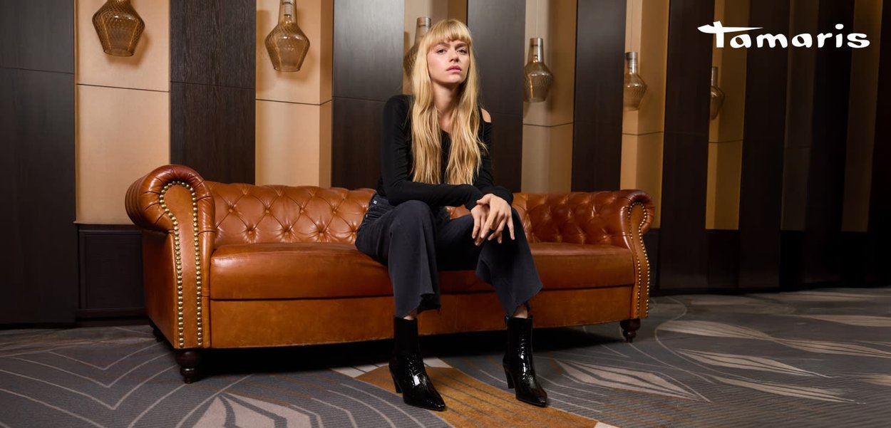 Canapé Chesterfield en cuir marron avec un design capitonné et des clous dorés. Modèle portant un haut noir et un pantalon foncé, avec des bottines noires brillantes.