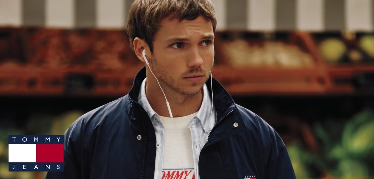 Joven con chaqueta azul marino y camiseta blanca de Tommy Jeans con auriculares, mirando ligeramente de lado en un ambiente interior.