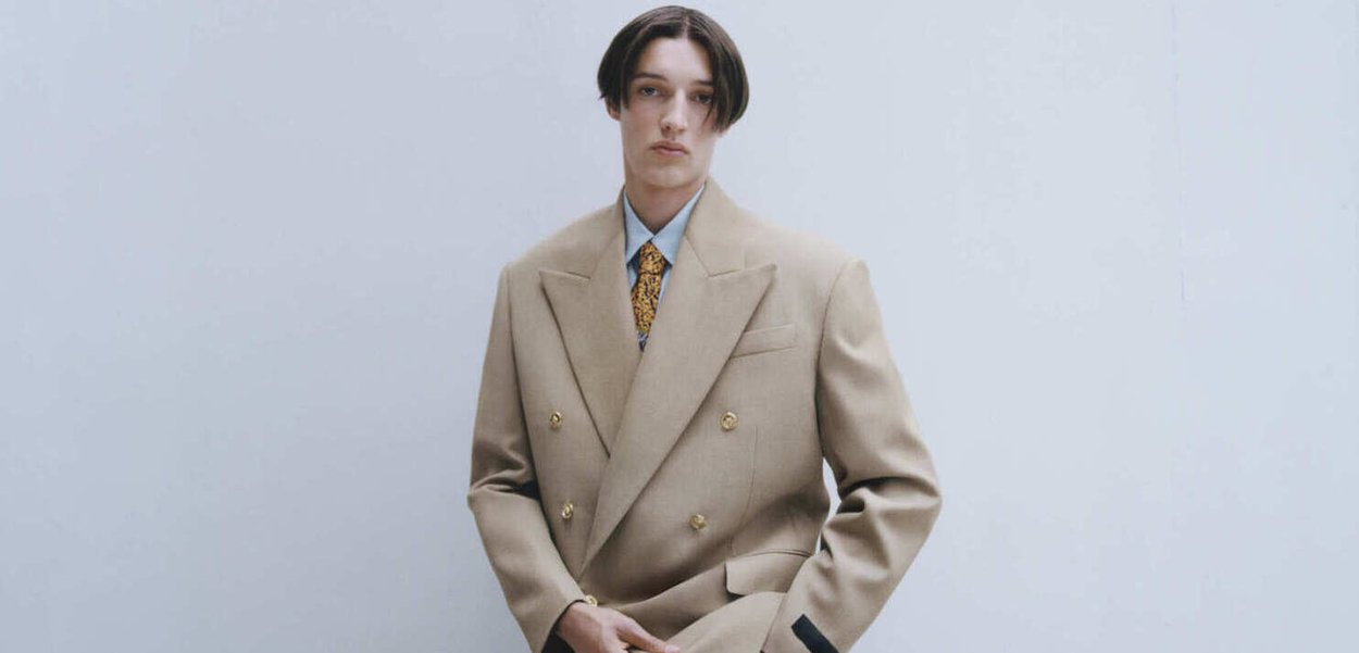Beige Doppelreiher-Sakko mit goldenen Knöpfen, kombiniert mit einem hellblauen Hemd und einer gemusterten Krawatte. Glatte Textur und taillierte Passform.