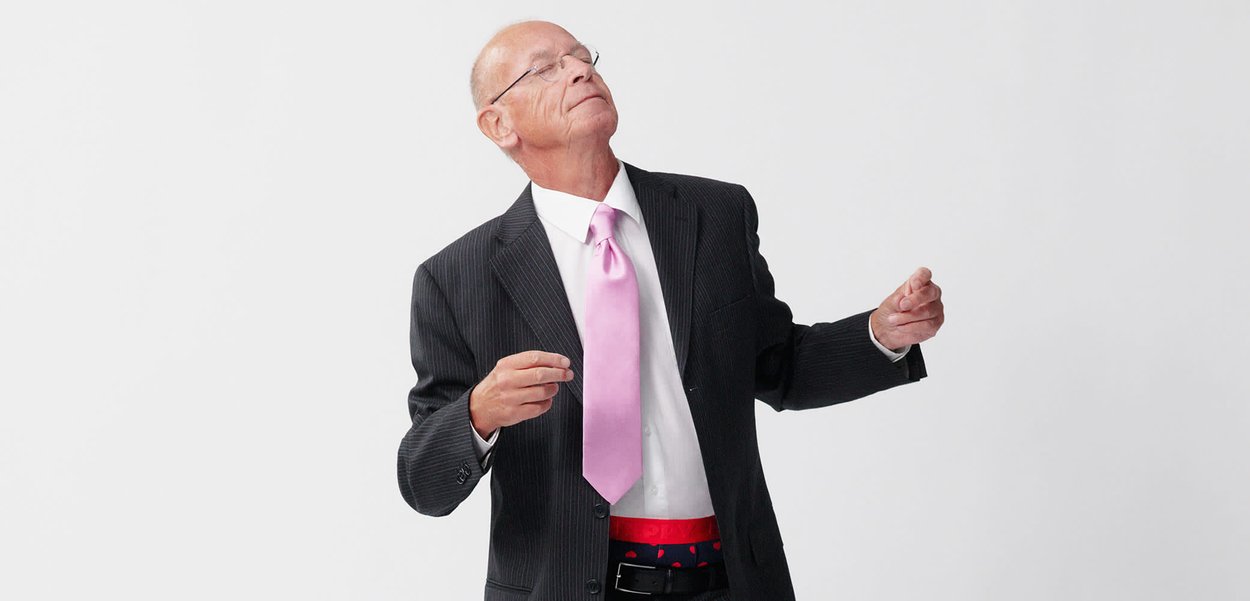 Schwarz gestreifter Anzug, weißes Hemd, rosa Krawatte, roter Gürtel mit Herzen, schwarze Schuhe. Stehende Pose, Arme leicht erhoben. Heller Hintergrund.