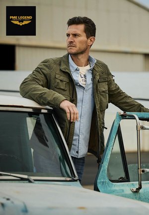 Hombre con chaqueta verde y camisa azul claro apoyado en la puerta abierta de un camión vintage azul claro desgastado al aire libre.