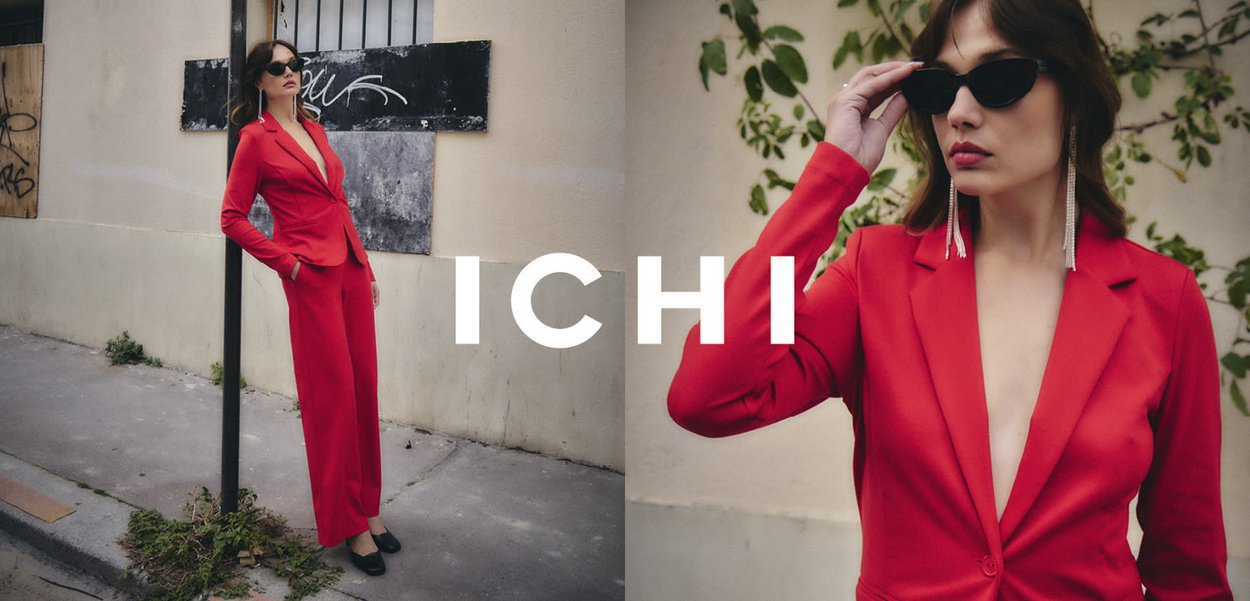 Roter, maßgeschneiderter Anzug mit tiefem V-Ausschnitt und langen Ärmeln. Accessoires umfassen übergroße Sonnenbrillen und baumelnde Ohrringe. Stadtkulisse.