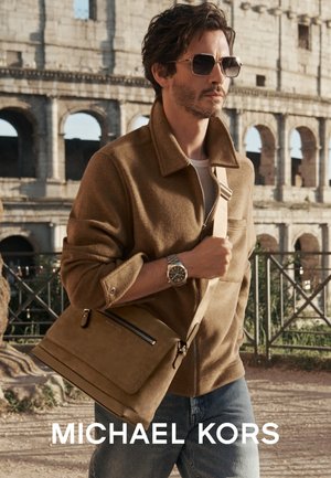 Veste en laine marron, sac bandoulière beige avec fermeture éclair, jean clair et montre en argent. En arrière-plan, le Colisée.