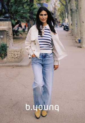 Eine Frau geht auf einem städtischen Bürgersteig und trägt eine beige Jacke, einen gestreiften Pullover, weit geschnittene Jeans und gelbe Schuhe. In der Nähe sind Bäume und Steinskulpturen zu sehen.