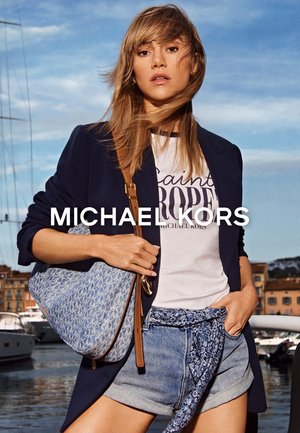 Joven mujer con blazer azul marino, camiseta blanca con logo, pantalones cortos de mezclilla con cinturón de pañuelo estampado y bolso azul con patrón, cerca de la marina.