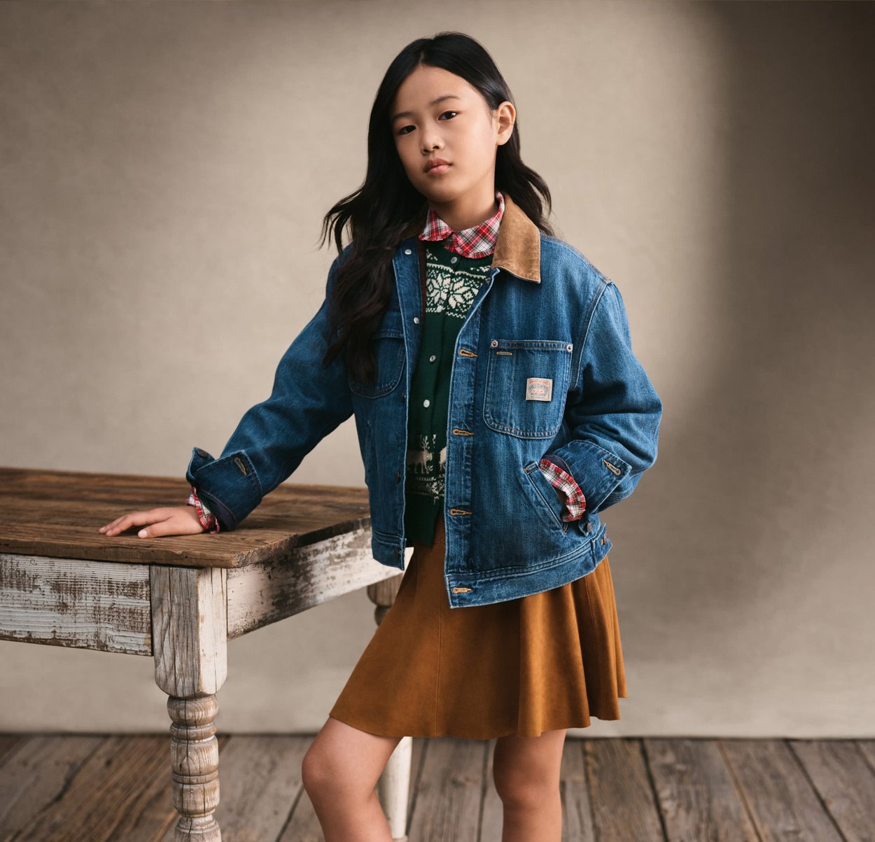 Ragazza giovane con capelli neri lunghi indossa giacca di jeans, maglione a motivo, camicia a quadri e gonna marrone, appoggiata a un tavolo rustico in legno.