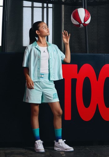 Ein Mädchen steht in einem hellblauen Jordan-Outfit, das aus einem kurzärmligen Shirt und Shorts besteht. Sie trägt weiße Sneaker und Socken mit Farbverlauf in Türkis und jongliert mit einem Basketball.