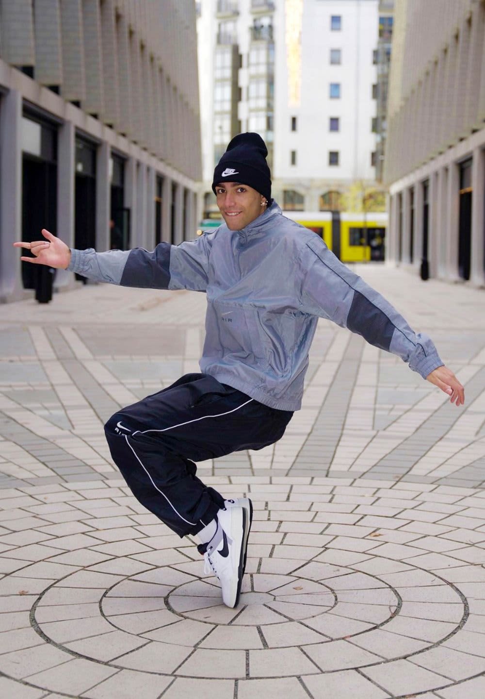 Gray windbreaker with black accents, black track pants with white stripes, and white sneakers. Standing on a circular patterned pavement.
