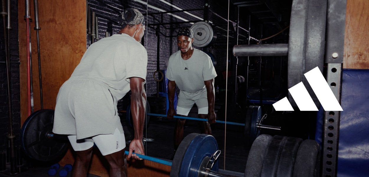 Homme soulevant une barre dans une salle de sport, portant des vêtements de sport légers et un bandeau, reflété dans un grand miroir avec des haltères et des équipements de gym autour.
