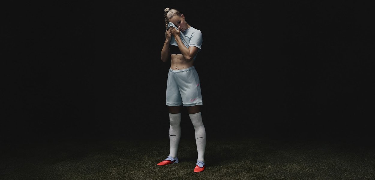 Lichtblauw sportuniform met een zwart crop top, voorzien van een Nike-logo. Rode voetbalschoenen en witte kniekousen. Staande pose.