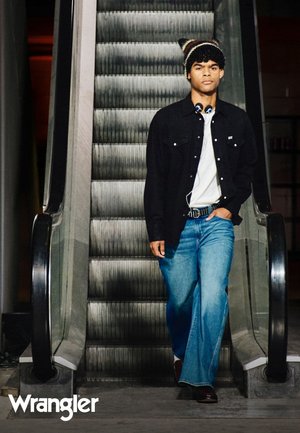 Modell med en svart denimjakke over en hvit skjorte, blå vide jeans og en mønstret ulllue, som står på en rulletrapp.