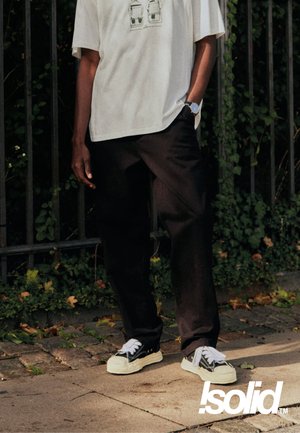 Person steht im Freien neben einem Metallzaun mit Grünpflanzen, trägt ein weißes T-Shirt mit Aufdruck, schwarze Hosen und schwarze Sneakers mit weißen Sohlen und Schnürsenkeln.