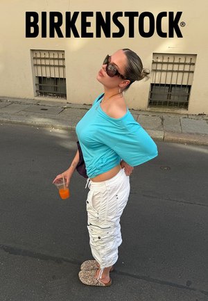 Woman wearing turquoise off-shoulder top, white cargo pants, leopard print Birkenstock sandals, holding orange drink, standing on street near wall with text.
