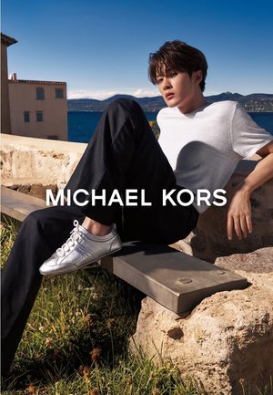 Ung mann i hvit t-skjorte og svarte bukser sitter på en steinbenk nær vannet, iført metalliske sølvfargede joggesko, med fjell i bakgrunnen.