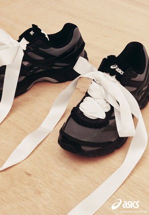 Par de zapatillas Asics negras y grises con cordones de cinta blanca extragrandes atados en grandes lazos sobre suelo de madera clara.