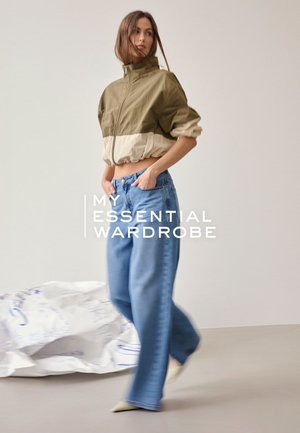 Frau trägt eine kurze beige-olivfarbene Jacke und hoch taillierte, lockere blaue Jeans, steht mit den Händen in den Taschen vor einem schlichten Hintergrund.