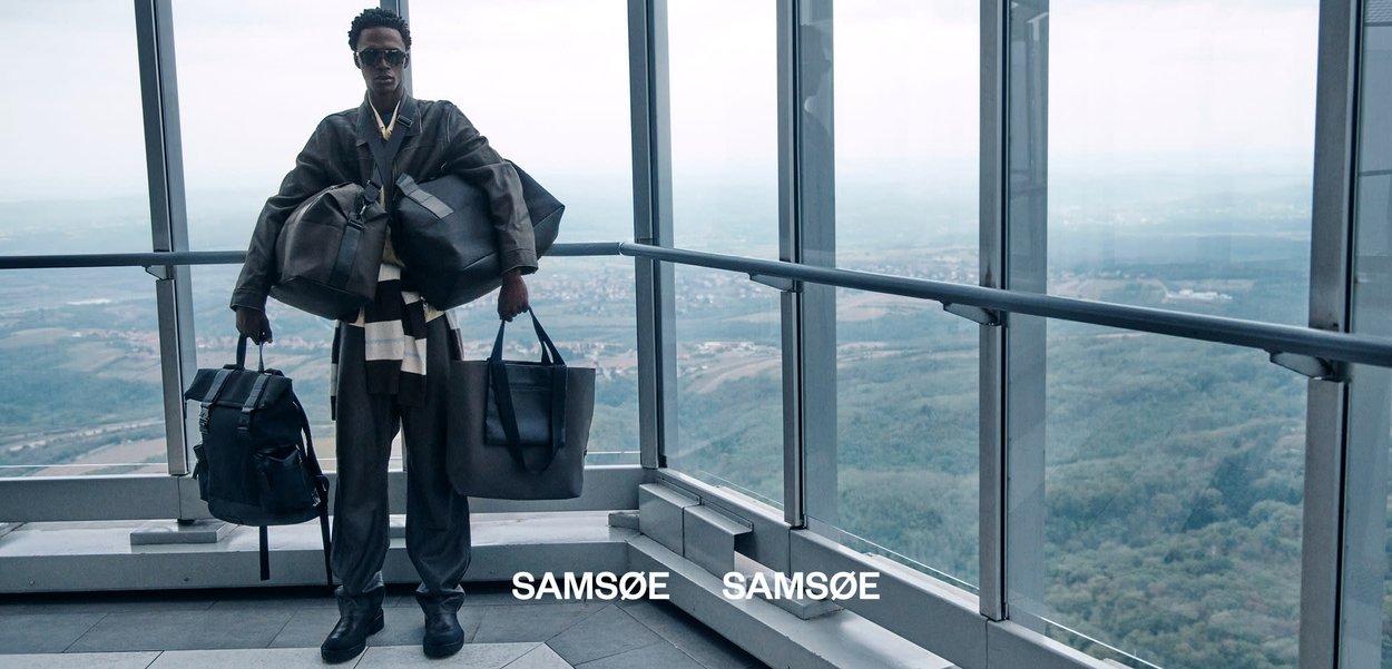 Mand iført oversized sort outfit, der holder tre store sorte tasker inde i en glaspudset højhus med udsigt over et landskab.