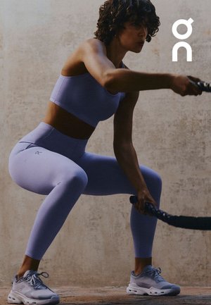 Mujer con ropa deportiva lila y zapatillas deportivas haciendo sentadillas mientras sostiene cuerdas de batalla contra una pared lisa con textura.