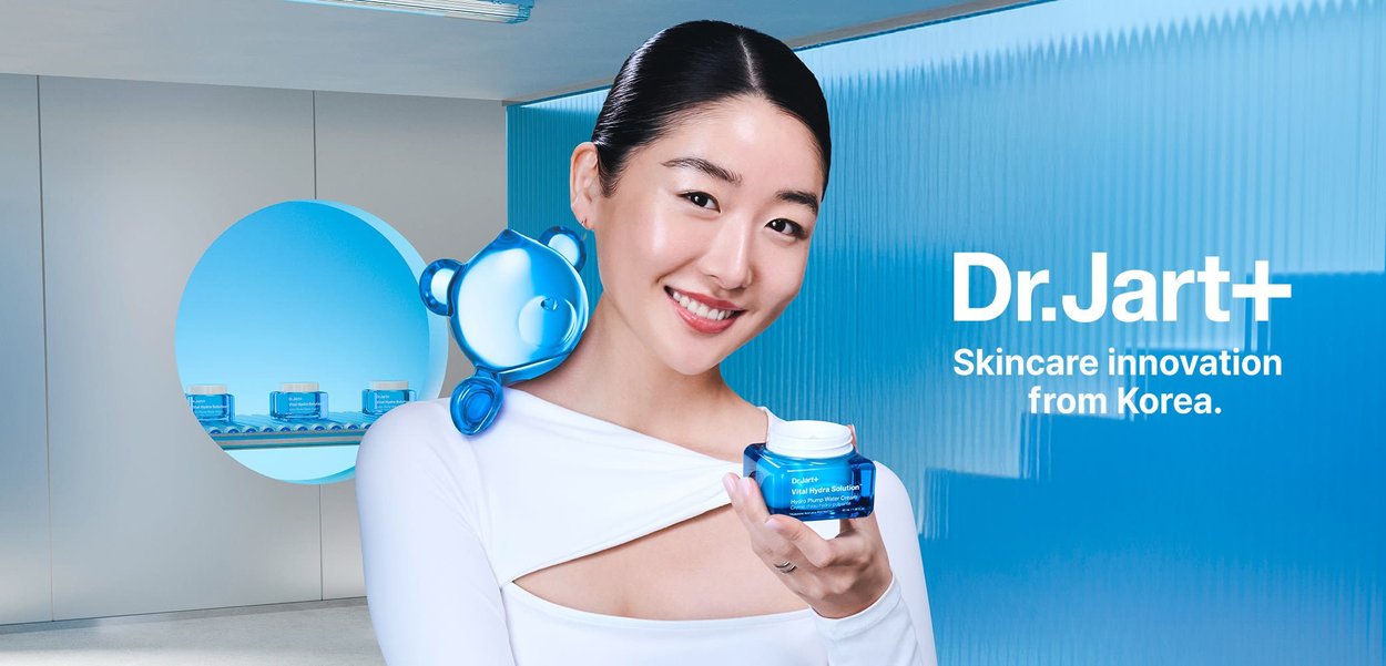 Dr. Jart+ Vital Hydra Solution cream in a blue jar, held by a person in a white top. Blue accents and a bear-shaped accessory on the shoulder.