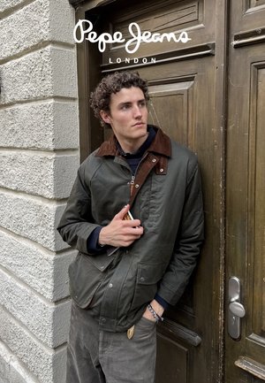 Chaqueta verde oscuro con un cuello de pana marrón, con cremallera y bolsillos, llevada sobre un suéter azul marino, sobre un fondo de puerta de madera.