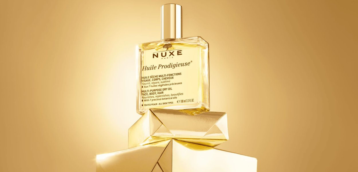 Bottiglia di vetro trasparente di olio secco NUXE con tappo argentato. Etichetta dorata con dettagli e testo. Esposizione su scatole geometriche dorate contro uno sfondo caldo.