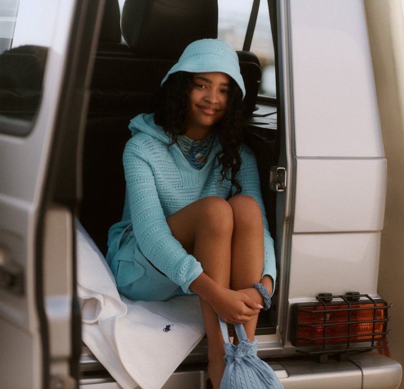 Joven sentada en la puerta lateral abierta de una furgoneta plateada, vestida con un suéter de punto azul claro y un sombrero tipo pescador, sosteniendo un bolso de punto a juego.