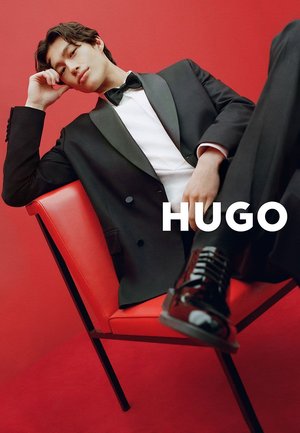 Schwarzer Smoking mit Satinrevers, weißem Hemd und Fliege, sitzend auf einem roten Stuhl vor rotem Hintergrund. Glänzende Schnürschuhe.