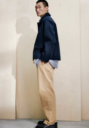 Homme debout contre un mur beige, portant une veste bleu marine, une chemise rayée, un pantalon beige et des chaussures noires, regardant de côté avec les mains dans les poches.