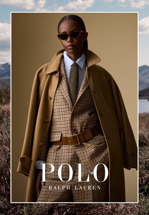 Ein Modell trägt einen beigen karierten Anzug mit brauem Gürtel, weißem Hemd, grauer Krawatte, einem über die Schultern drapierten Kamelfarbenen Mantel und einer Brille mit Schildpattmuster.