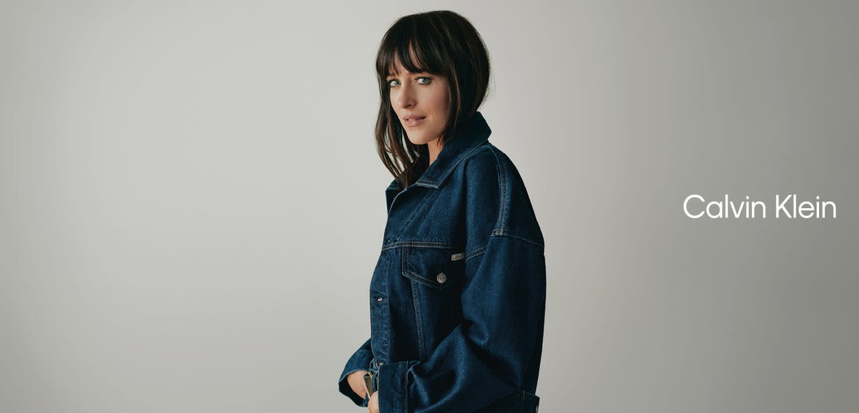 Mujer de cabello oscuro con una chaqueta vaquera azul oscura oversize, de pie contra un fondo claro y liso junto al logotipo de Calvin Klein.