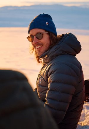 Gepolsterte graue Jacke mit hohem Kragen, blauer Strickmütze und Sonnenbrille. Natürliche Landschaft im Hintergrund mit sanften Sonnenuntergangsfarben.