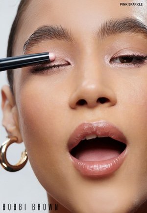 Crayon pour les yeux Bobbi Brown en rose vibrant scintillant appliqué sur la paupière ; le modèle a des sourcils foncés, une peau claire et des lèvres nude brillantes, avec des boucles d'oreilles en or.