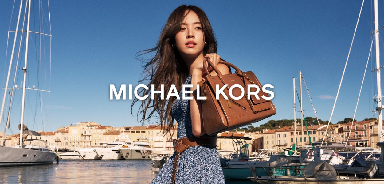 Ung kvinne i blå kjole holder en brun skinnveske nær en marina med yachter under en klar blå himmel.