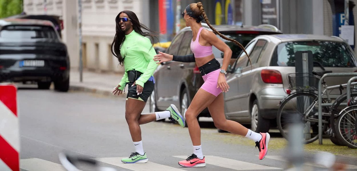 Dos mujeres vestidas con ropa deportiva corren alegremente por un paso de peatones en una calle de la ciudad con coches y bicicletas aparcados cerca.