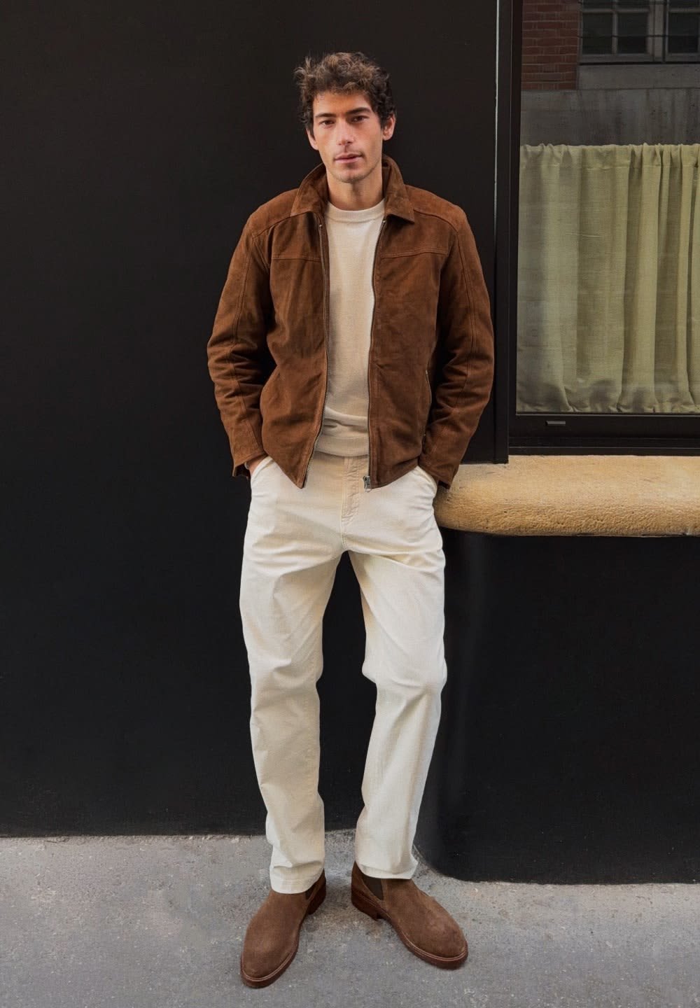 Bruine suède jas over een lichte trui, crèmekleurige broek en bruine enkellaarsjes. Effen kleuren en eenvoudig ontwerp, staand tegen een zwarte muur.