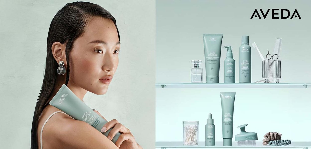 Aveda haarverzorgingslijn op een plank; lichtgroene tubes en flessen, glazen containers en metalen gereedschap. Zachte textuur.