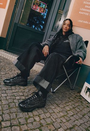 Baskets New Balance noires avec une finition brillante, présentant des détails en mesh et en cuir. Assorties à un pantalon large noir et une veste bomber grise.