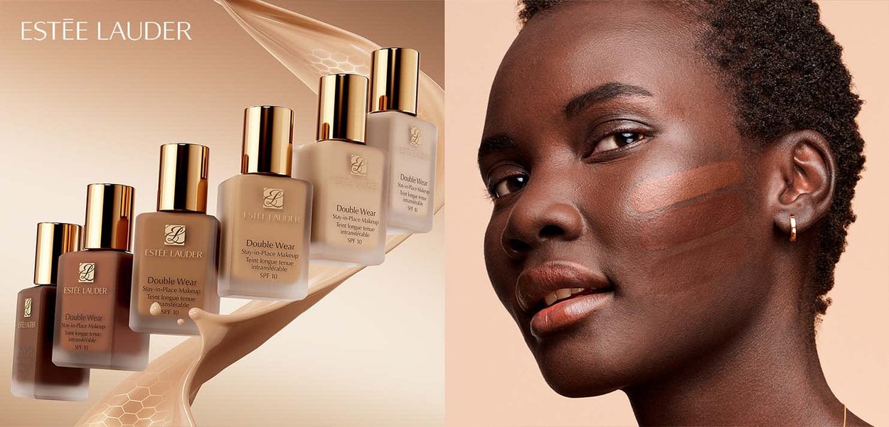 Donna dalla pelle scura con campioni di fondotinta sulla guancia accanto a sei bottiglie di fondotinta Estee Lauder Double Wear in diverse tonalità.