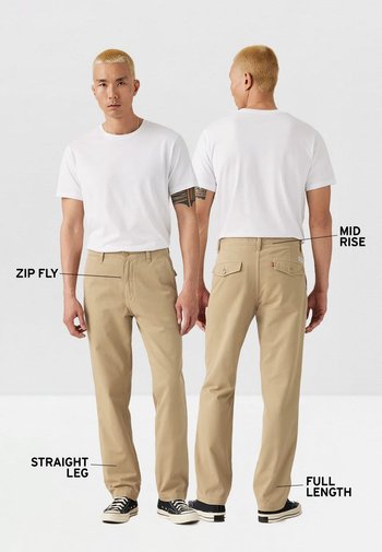 Pantalones beige de pierna recta con cremallera, cintura de altura media y longitud completa. El modelo lleva una camiseta blanca y zapatillas negras.