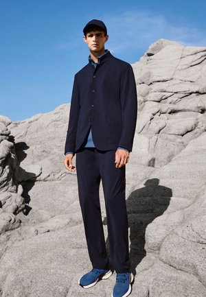 Giovane uomo si trova su un terreno roccioso indossando un completo blu navy, scarpe da ginnastica blu e un berretto da baseball abbinato, sotto un cielo azzurro limpido.