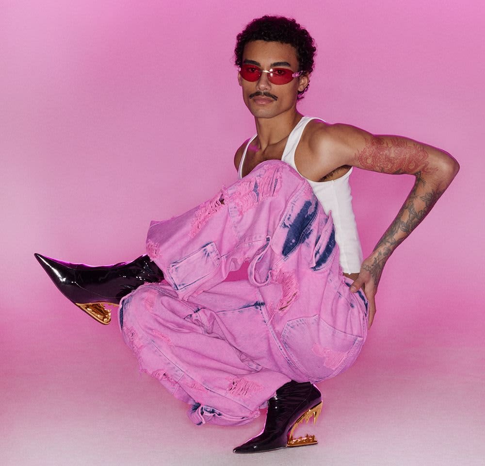 Pink distressed baggy jeans with a loose fit, paired with a white tank top and glossy black boots featuring gold accents. Red-tinted sunglasses.