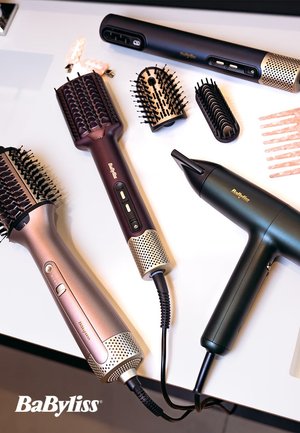 Quatre outils de coiffure sur une surface blanche : un sèche-cheveux noir, un sèche-cheveux brosse rond rose doré, et deux petites brosses de couleurs sombres, toutes avec différentes textures.